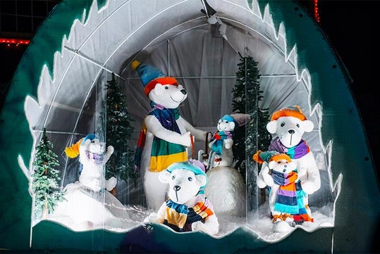 Five polar bear figures wearing colorful winter clothes are displayed in a snowy, decorated enclosure with artificial trees and a snowman in the background.