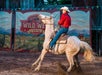 A man in a red shirt and white hat riding a white speckled horse and pulling on the reigs.