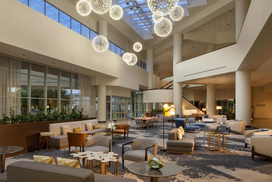 Spacious hotel lobby with modern furniture, tall columns, and decorative lighting. Large windows provide natural light.