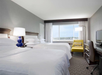 Hotel room with two large beds, white linens, a yellow chair, desk, and TV. Large window with a view. Gray carpet and walls.