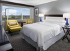 Hotel room with a large bed, two yellow chairs, and a round table. A window shows a view of a theme park with roller coasters.