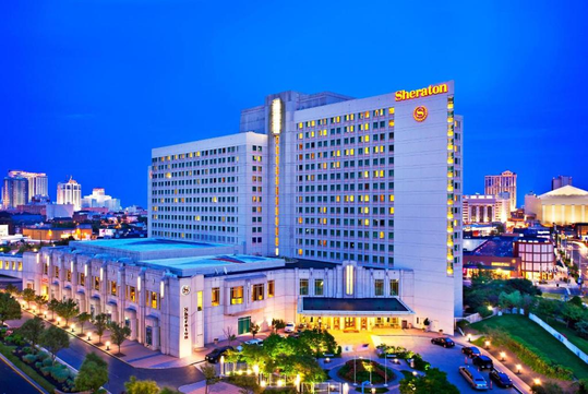 A large hotel with the sign "Sheraton" is illuminated at night against the city skyline.