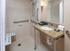 Bathroom with a walk-in shower, grab bars, and a foldable seat. Granite countertop with a sink, mirror, towels, and toiletries. Beige tiles on walls and floor.