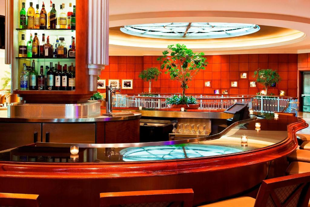 A modern bar with a marble counter and a selection of bottles. Candles are lit on the counter, and there are potted plants and a glass ceiling in the background.