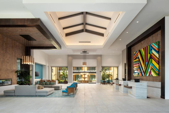 Modern hotel lobby with high ceiling, geometric artwork on the wall, several seating areas with blue and beige sofas, and a reception desk on the right.