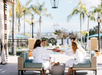 Four people in white robes sit on outdoor sofas, toasting drinks under a canopy, with palm trees and a pool in the background.