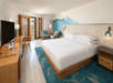 Hotel room with a king-sized bed, wall art, TV, and a balcony with a view. Room includes a desk, armchairs, and ambient lighting.