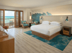 Hotel room with a large bed, two chairs, and a view of the ocean through the open balcony doors. The decor features a wave mural and turquoise accents.