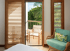 A cozy room with wooden shutters, a turquoise chair with a floral cushion, and an open door leading to a balcony with two chairs and lush greenery outside.
