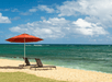 Two lounge chairs under a red umbrella on a sandy beach by the ocean, with a partly cloudy sky overhead.