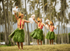Three dancers in traditional Hawaiian outfits perform outdoors, wearing grass skirts and floral leis, with palm trees in the background.