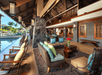 A luxurious open-air resort lounge featuring wicker furniture with colorful cushions, a stone wall, and lantern lighting overlooking a pool and palm trees.