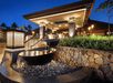 Water flows from two large bowls in a modern outdoor fountain, surrounded by stone walls and lush greenery, at night. A building with warm lighting and steps is visible in the background.