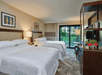 Hotel room with two double beds, a TV on a stand, and a patio door opening to a garden view. Lamps and framed artwork adorn the walls.