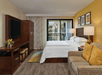 Hotel room with a bed, beige sofa, and wooden furniture. A flat-screen TV is on a cabinet. Sliding doors open to a balcony. Walls are painted yellow and beige, with framed artwork above the bed.