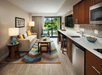 Modern hotel room with a kitchenette, seating area featuring a beige sofa, vibrant chevron-patterned rug, and large sliding glass doors opening to a garden view.