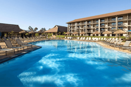 A large outdoor swimming pool surrounded by deck chairs, with a multi-story hotel in the background under a clear blue sky.