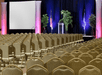Empty conference room with rows of beige chairs facing a stage equipped with a podium and large projection screen, flanked by potted plants and flags, under colorful lighting.