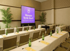 Conference room with rows of tables and chairs, each table set with green bottles and yellow notepads. A screen displays "Sheraton Myrtle Beach Convention Center Hotel.