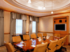 A modern conference room with a long wooden table surrounded by beige chairs, with a large wall-mounted screen and curtained windows.