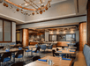 Modern restaurant interior with wooden tables, blue chairs, and an open kitchen. A chandelier hangs from the ceiling. Blue napkins and glassware are neatly arranged on the tables.