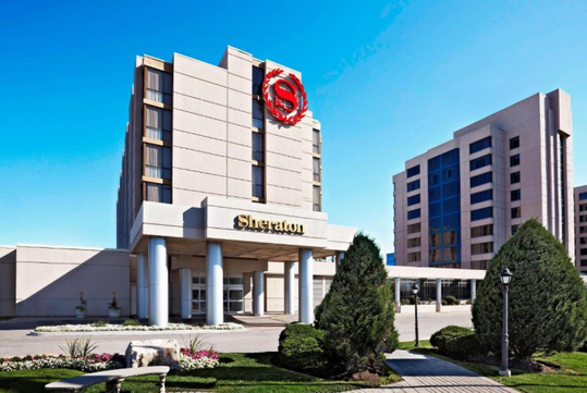 Hotel exterior with a prominent red logo on a building facade, flanked by a grassy area and trees under a clear blue sky.