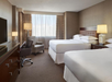 A hotel room with two double beds, a modern chair, a desk with a chair, a TV on a dresser, and a window with curtains.