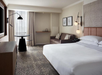 Hotel room with a large bed, a sofa, a round table, and a wall-mounted TV. Minimalist decor includes framed artwork and contemporary lighting.