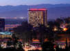 A tall hotel building with "Sheraton Universal" signage is illuminated at dusk, surrounded by trees and city lights, with mountains visible in the background.