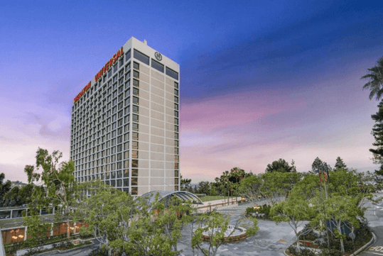 A tall hotel building labeled "Sheraton Universal" is surrounded by trees and landscaping under a purple and pink sky at dusk.