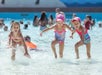 Three young girls in swimsuits and pink caps play and splash in a crowded shallow pool, with other people visible in the background.