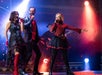 Three singers perform on stage under colorful lights, wearing black and red outfits, with a musician playing guitar in the background.