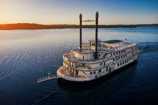 Paddle steamboat with two tall smokestacks on calm waters at sunset, with distant shoreline in the background.
