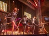 Performers in sparkly outfits dance energetically on a brightly lit stage with musicians in the background.