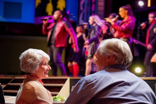 An elderly couple smiles at each other while sitting at a table. A band performs energetically on stage in the background.