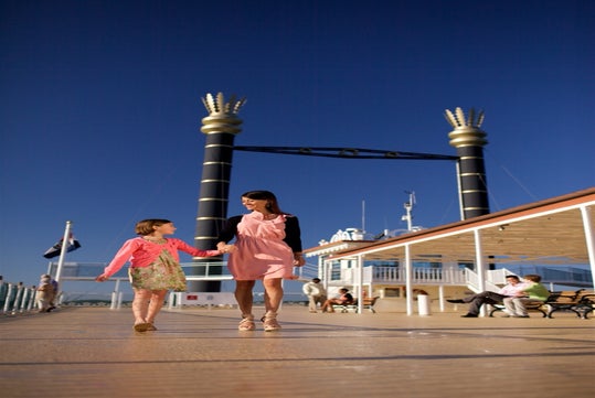 A woman and a young girl walk hand-in-hand on the deck of a cruise ship under a clear blue sky, with other passengers seated in the background.