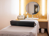 A massage room with a bed, a rolled towel, a large round mirror, a sink, and soft lighting against a beige wall.