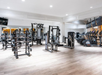 A spacious gym interior with various exercise machines, including treadmills, weight racks, and other fitness equipment, on a light wooden floor. Large mirrors line the walls.