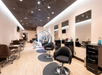 A modern hair salon with styling chairs, mirrors, and workstations lining the walls. The interior features a clean, minimalistic design with light wood floors and overhead lighting.