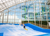 Person surfing on an indoor wave machine inside a large glass structure with visible slides in the background.