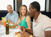 Three people sit at a dining table, eating and conversing. Plates of food and drinks are in front of them.