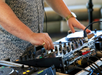 A person in a patterned shirt adjusts controls on a DJ mixer, surrounded by audio equipment.