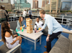 Four people are playing Jenga on a boat deck, surrounded by city buildings and water.