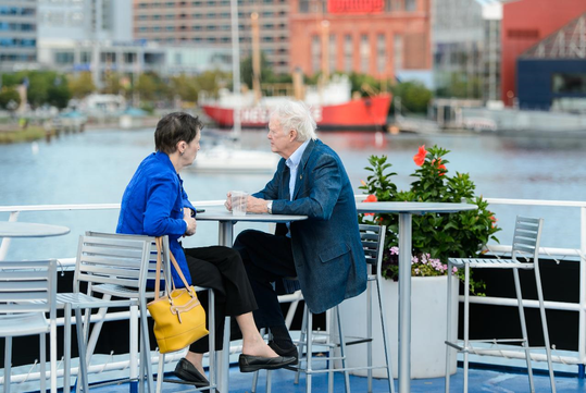 Two people sit at a table on a waterfront deck, talking and drinking coffee. A red ship and buildings are visible in the background.