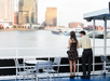 Two people stand on a deck overlooking a city skyline and water with boats. Metal chairs and tables are visible in the foreground.