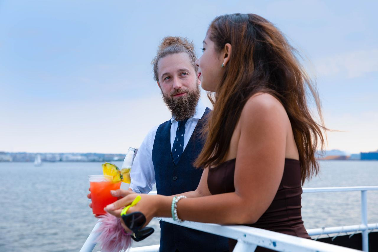 A man and woman chat on a boat deck, holding drinks. The water and distant coastline are visible in the background.