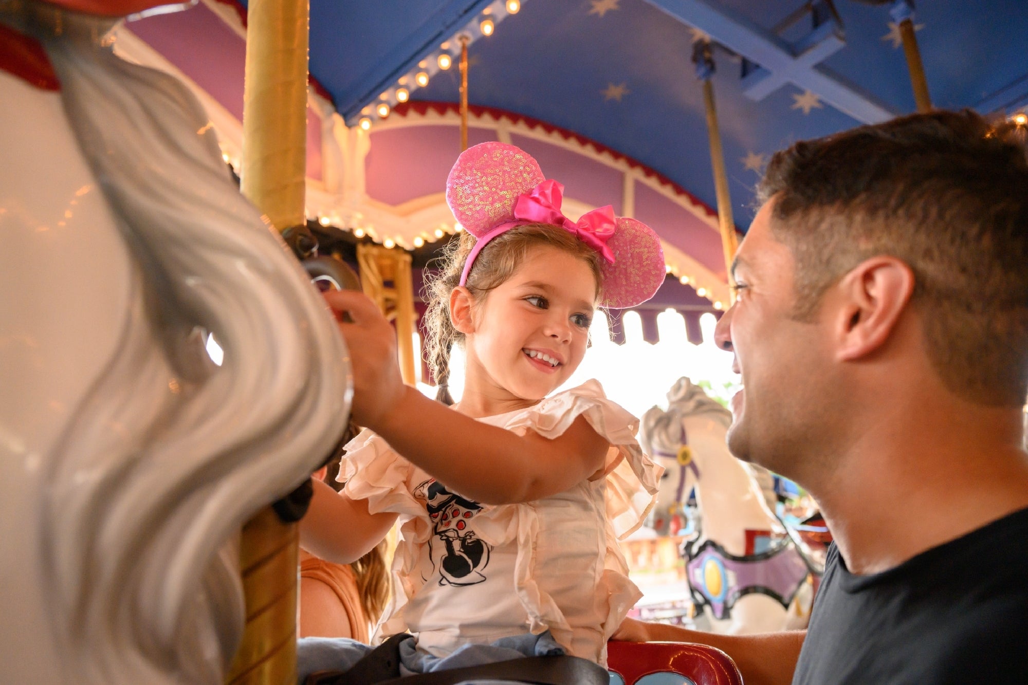 A young girl wearing pink mouse ears smiles at an adult while riding a carousel with brightly colored horses in the background.