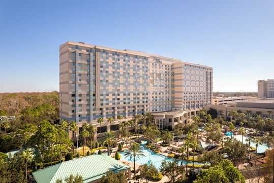 A large, modern hotel building with many windows stands next to a landscaped pool area surrounded by palm trees under a clear blue sky.