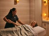 A massage therapist gives an arm massage to a person lying on a spa table, covered with a gray blanket, in a softly lit room.
