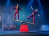 Two circus performers in matching costumes balance on opposite ends of a wooden board placed on a rolling cylinder, with flags and barrels in the background under stage lights.